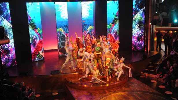Eine Gruppe von Darstellern in farbenfrohen Kostümen auf einer Bühne vor einem Unterwasser-Hintergrund.
