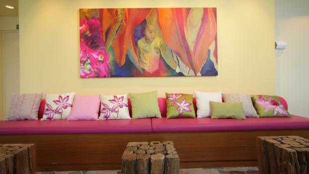 Ein Wartezimmer mit einer pinkfarbenen Bank, vielen Kissen und einem bunten Gemälde an der Wand.