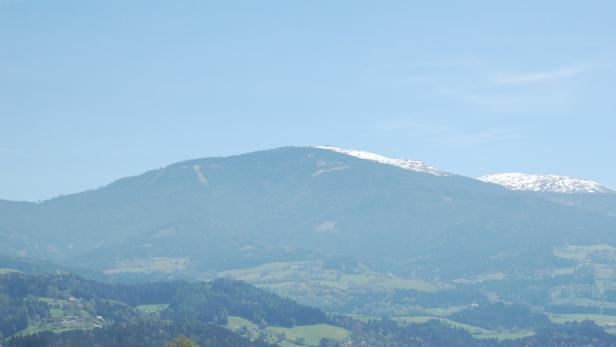 Hügelige Landschaft mit bewaldeten Hügeln und schneebedeckten Gipfeln unter blauem Himmel.