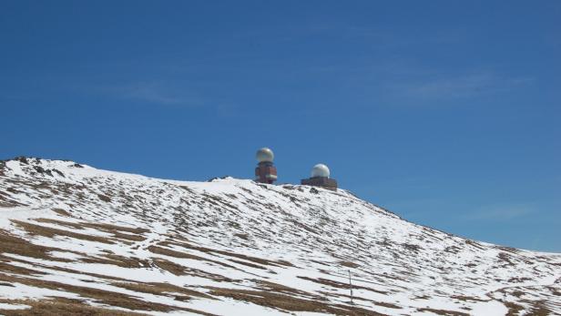 Zwei Gebäude mit Radarkuppeln auf einem schneebedeckten Hügel unter blauem Himmel.