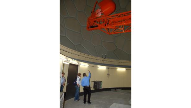 Besucher stehen unter der Kuppel eines Observatoriums und betrachten ein orangefarbenes Teleskop.