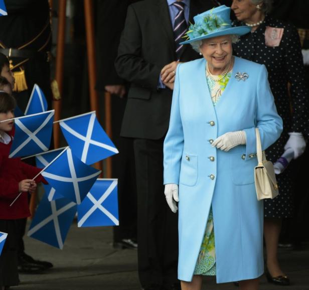 Königin Elisabeth II. in einem hellblauen Mantel, umgeben von schottischen Flaggen.