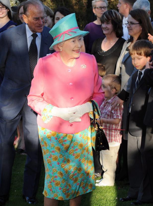 Königin Elisabeth II. trägt einen pinkfarbenen Blazer und einen türkisfarbenen Hut.