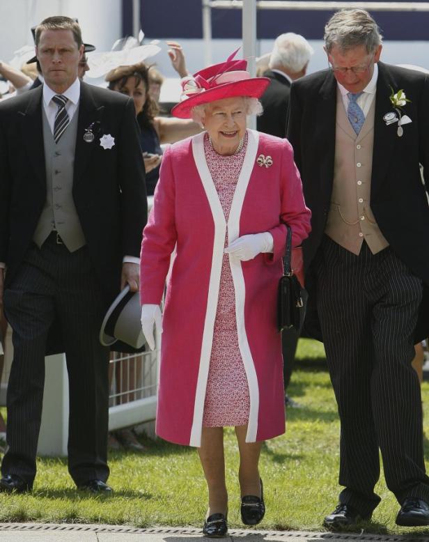 Königin Elisabeth II. in einem pinkfarbenen Mantel und Hut, begleitet von zwei Männern.