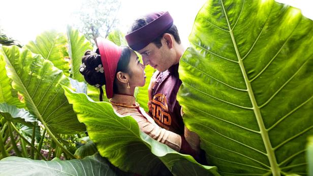 Ein Paar in traditioneller vietnamesischer Kleidung steht inmitten großer grüner Blätter.