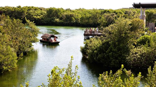 Ein Boot mit Strohdach fährt auf einem Fluss in Richtung eines Holzstegs mit Personen.