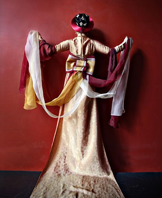 Eine Person in einem traditionellen chinesischen Kleid mit weiten Ärmeln vor einem roten Hintergrund.