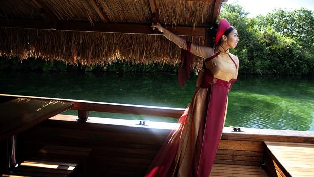 Eine Frau in traditioneller Kleidung blickt von einem Boot auf einen grünen Fluss.