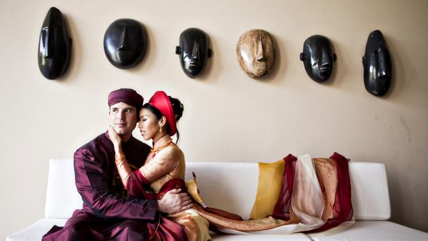Ein Paar in traditioneller vietnamesischer Kleidung sitzt vor einer Wand mit Masken.