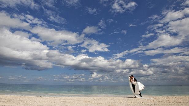Ein Bräutigam trägt seine Braut am Strand unter einem bewölkten Himmel.
