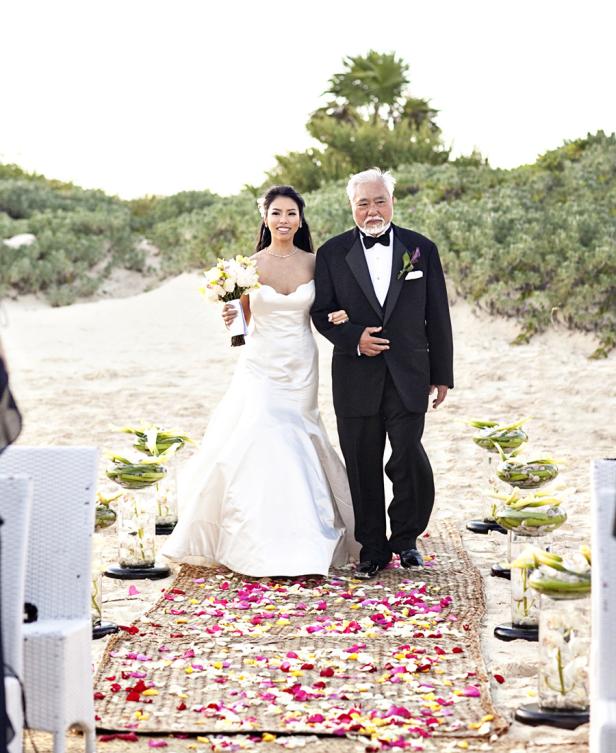 Eine Braut wird von ihrem Vater am Strand entlang geführt.