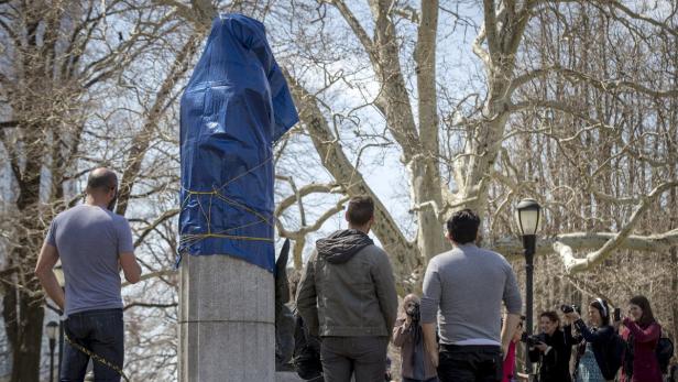 Eine mit einer blauen Plane verhüllte Statue wird von mehreren Personen fotografiert.