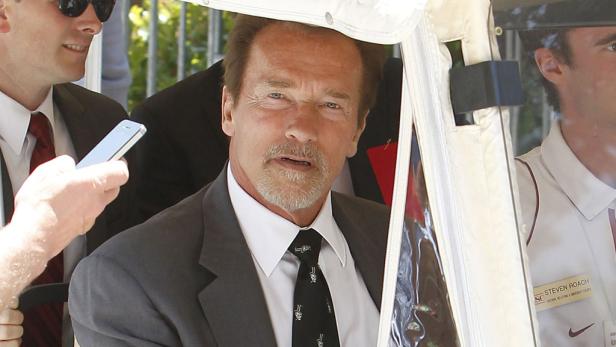Arnold Schwarzenegger sitzt in einem Golfwagen und wird von Menschen umringt.