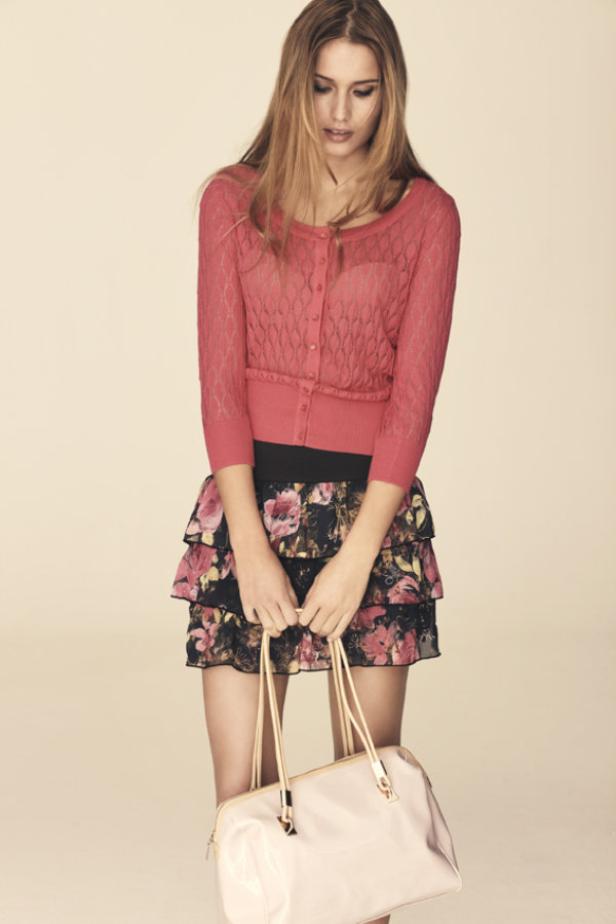 Eine junge Frau mit einem rosa Cardigan und einem geblümten Rock hält eine Handtasche.