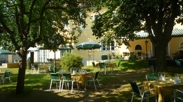Ein Restaurantgarten mit Tischen und Stühlen unter schattenspendenden Bäumen.