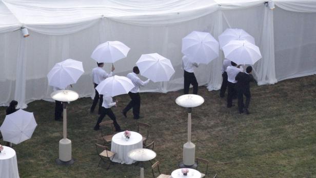 Mehrere Personen mit weißen Hemden und Regenschirmen gehen über einen Rasen vor einem weißen Zelt.