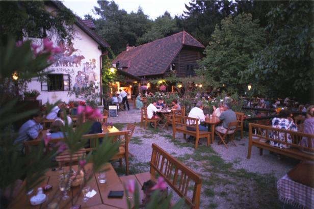 Ein Biergarten mit vielen Gästen an Tischen unter Bäumen.