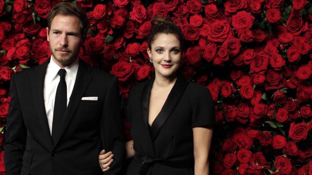 Drew Barrymore posiert mit einem Mann vor einer Wand aus roten Rosen.