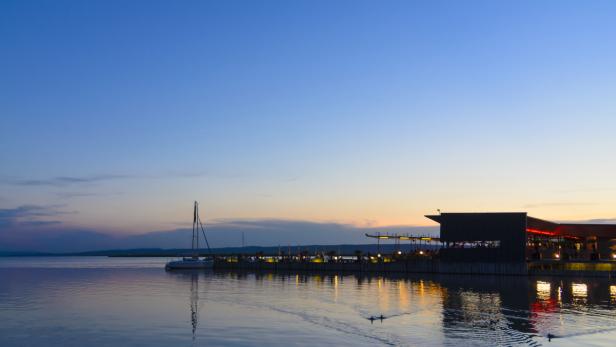 Ein See mit einem Restaurant auf einem Pier und einem Segelboot im Abendlicht.