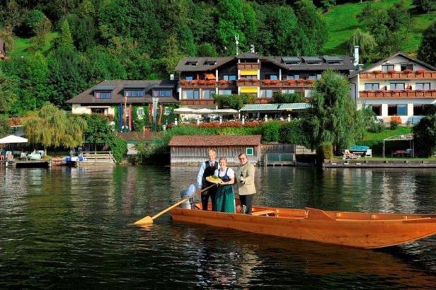 Drei Personen in Tracht fahren mit einem Holzboot auf einem See an einem Hotel vorbei.