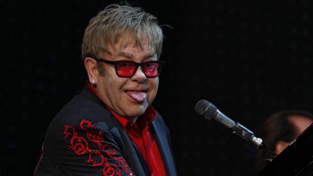 Elton John streckt die Zunge heraus, während er am Klavier sitzt.
