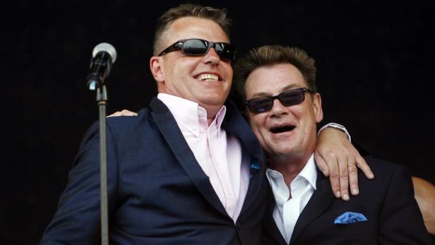 Zwei Männer in Anzügen und Sonnenbrillen stehen lächelnd auf einer Bühne vor einem Mikrofon.