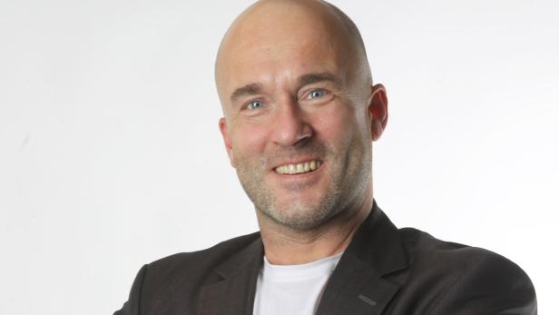 Ein Mann mit Glatze lächelt in einem dunklen Sakko und einem weißen T-Shirt.