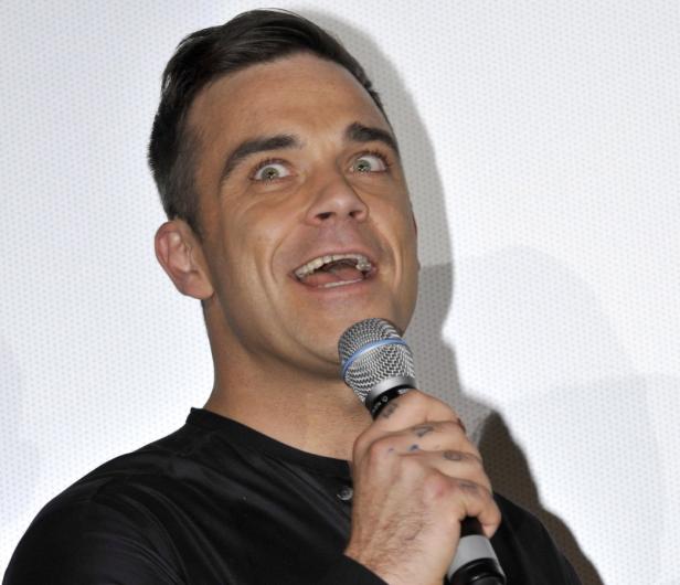Robbie Williams spricht in ein Mikrofon und blickt mit offenem Mund nach oben.