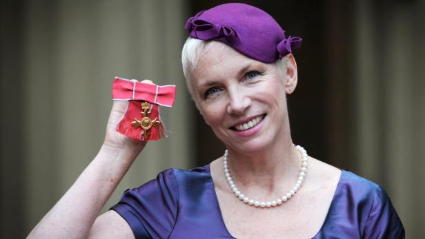 Annie Lennox mit einem lila Hut und einer Halskette hält einen britischen Verdienstorden in der Hand.
