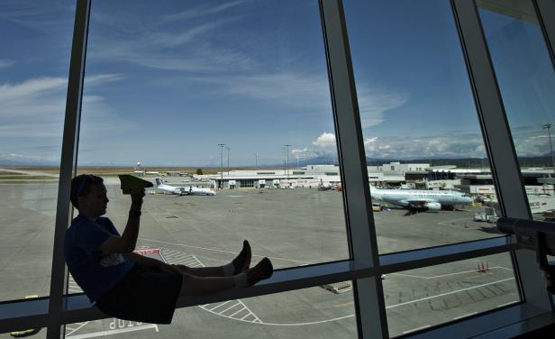 Ein Junge sitzt am Fenster eines Flughafens und hält ein Papierflugzeug in der Hand.