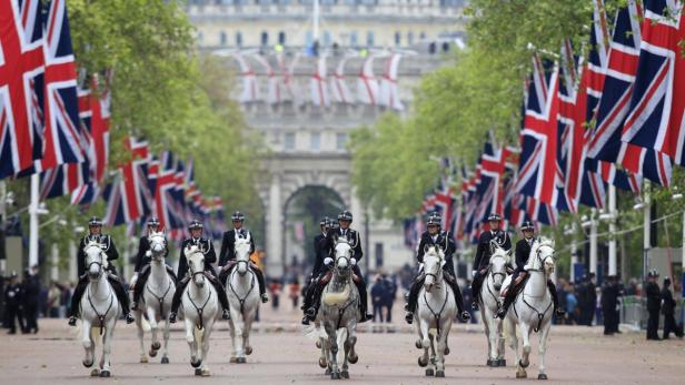 Polizisten auf weißen Pferden reiten eine mit britischen Flaggen geschmückte Straße entlang.