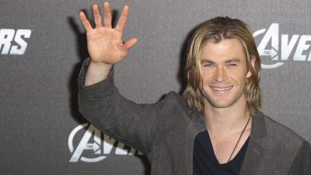Chris Hemsworth lächelt und winkt vor einem „Avengers“-Logo.