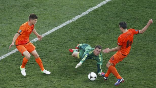 Ein Fußballspiel mit einem Torwart in Grün, der versucht, den Ball abzuwehren, während zwei Spieler in Orange zusehen.