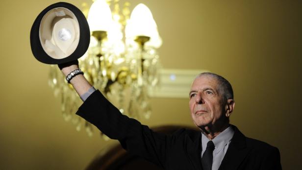 Leonard Cohen hält einen Hut in die Höhe.
