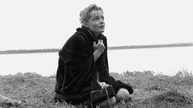 Eine junge Frau sitzt in einen Bademantel gehüllt am Ufer eines Sees.