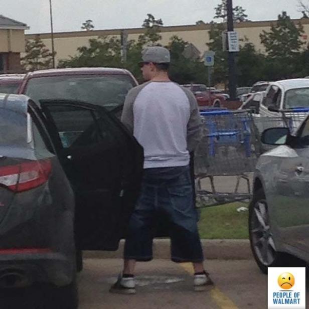 Ein Mann steht auf einem Parkplatz neben einem Auto und trägt seltsame Jeansshorts.