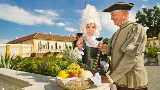 Zwei Personen in historischen Kostümen halten Weingläser vor einem Schloss und einem Obstkorb.