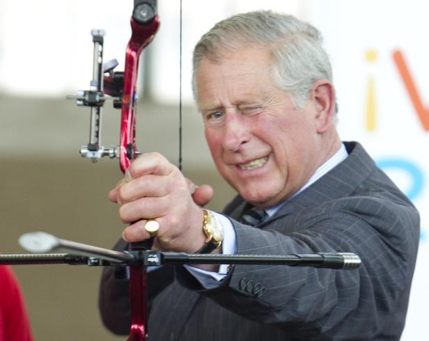 König Charles zielt mit einem roten Bogen und zwinkert dabei.