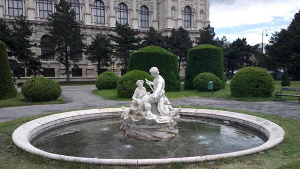 Ein Brunnen mit einer Skulpturengruppe im Park vor einem Gebäude in Wien.