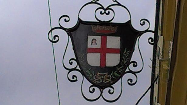 Ein verziertes Schild mit einem Wappen hängt an einem Gebäude.