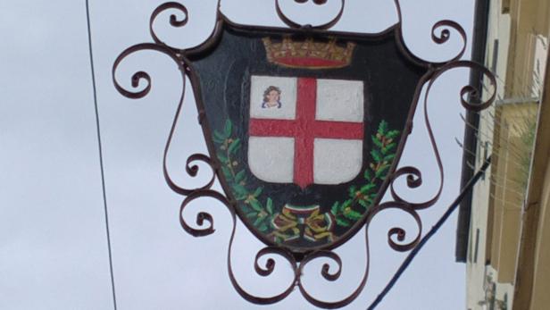 Ein Wappen mit einem roten Kreuz, einer Krone und einer kleinen Figur an einer verzierten Halterung.