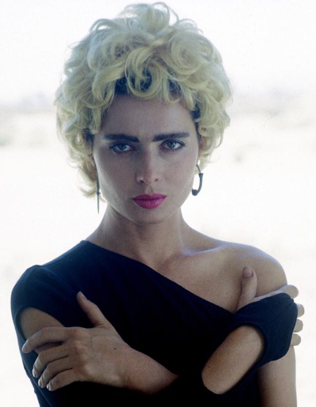 Eine Frau mit blond gelocktem Haar und dunklem Kleid blickt in die Kamera.