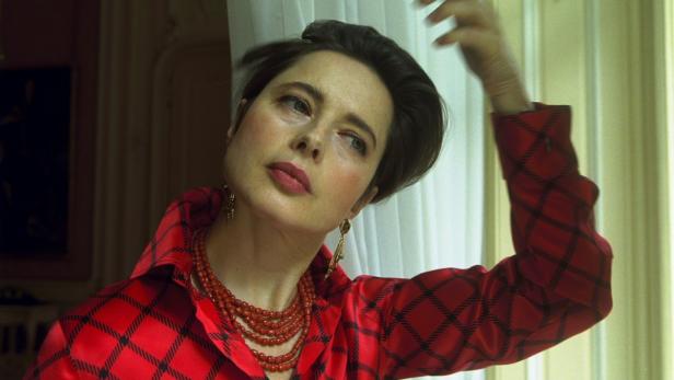 Eine Frau mit kurzem, dunklem Haar trägt eine rote Bluse mit Karomuster und eine rote Perlenkette.