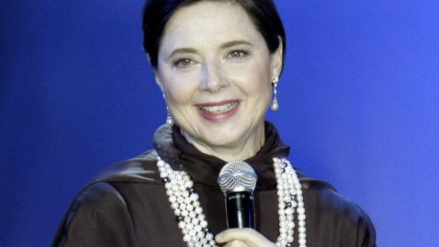 Eine Frau mit Perlenkette hält ein Mikrofon vor einem blauen Hintergrund.