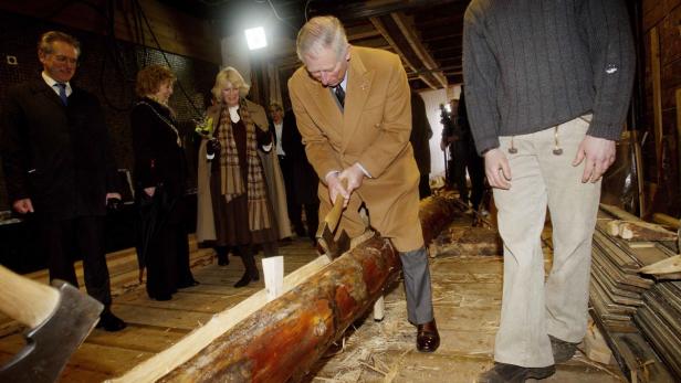 König Charles hackt mit einer Axt einen Baumstamm, während Camilla und andere zusehen.