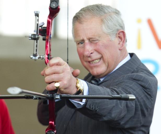 König Charles zielt mit einem roten Bogen und zwinkert dabei.
