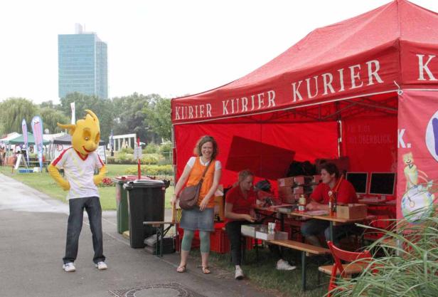 Ein gelbes Maskottchen steht vor einem roten „Kurier“-Zelt, in dem Leute sitzen.