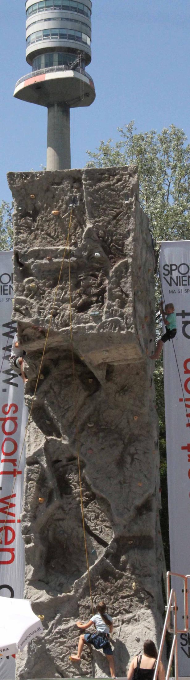 Mehrere Personen klettern an einem künstlichen Felsen vor dem Hintergrund des Donauturms.