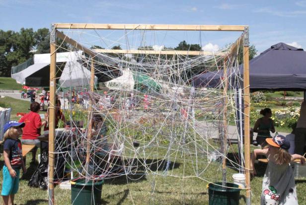 Ein Holzrahmen ist mit vielen bunten Schnüren umwickelt, umgeben von Menschen in einem Park.