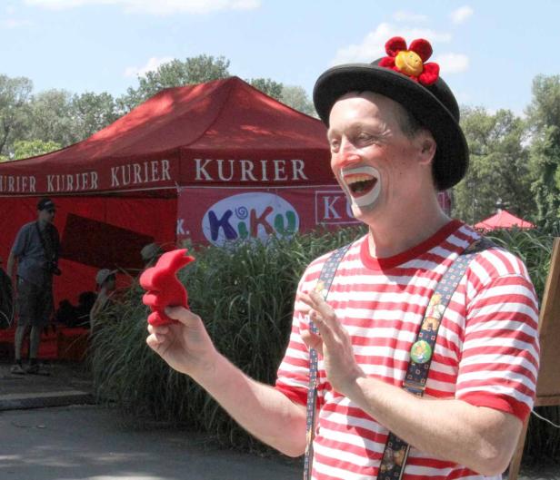 Ein lachender Clown mit roter Nase und Hut hält eine rote Hasenfigur in der Hand.